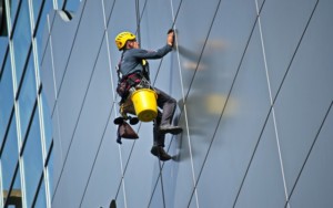 A worker wearing a helmet and safety harness performs deep track cleaning while cleaning the exterior glass windows of a tall building using suspended equipment.