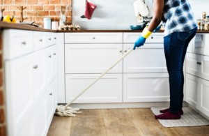 Person wearing gloves mops the kitchen floor near white cabinets, standing on a mat in front of a countertop with various kitchen items, tackling hard water staining for a spotless finish.