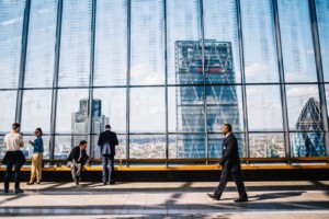 People in business attire stand and walk inside a modern glass building, with the city skyline and high-rise buildings visible through large windows adorned with subtly scratched glass.