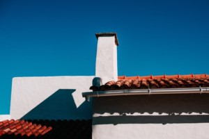A white building with a chimney and red tiled roof is shown against a clear blue sky, while a gutter technician cleaning keeps the exterior pristine.