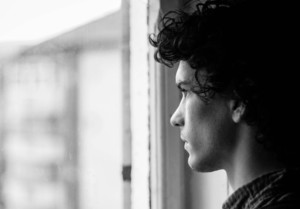 A young person with curly hair looks out of a window marked by faint water stains. The black and white image captures the side profile of their face, adding a contemplative mood to the scene.