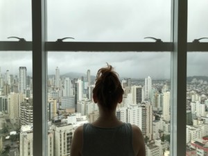 Person with hair in a bun stands indoors facing large windows, looking out at a city skyline with many tall buildings under a cloudy sky, as subtle hard water staining marks the glass.