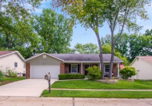 Single-story brick house with attached garage, green lawn treated with sodium hypochlorite, trees in front yard, and an American flag by the entrance on a suburban street.