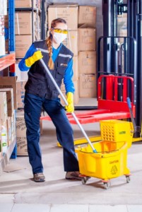 A person wearing safety glasses and gloves is mopping the floor in a warehouse, next to a yellow caution mop bucket, stacked cardboard boxes, and equipment used for traditional window cleaning.