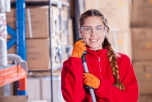 A woman wearing safety glasses, orange work gloves, and a red jacket smiles while holding a mop used for traditional window cleaning in a warehouse with cardboard boxes in the background.