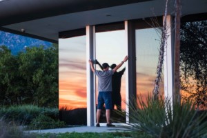 A person stands at a large glass door of a modern building, holding it open, with a sunset and trees reflected in the freshly cleaned glass—hinting at recent time window cleaning.