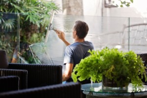 A person uses a squeegee to clean a glass panel in an outdoor area with wicker furniture and green plants, illustrating the quality you can expect when considering professional window cleaning pricing.