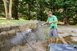 A man wearing sunglasses uses a pressure washer to clean a stone retaining wall in an outdoor Kelowna yard surrounded by trees.