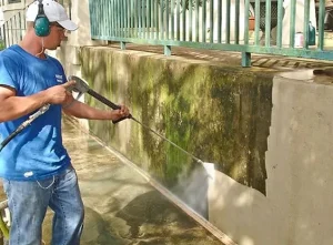 A man uses a pressure washer for house washing in Kelowna, removing moss and dirt from a concrete wall to reveal a lighter, clean surface beneath.