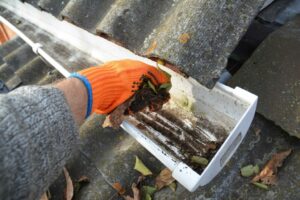 A person wearing an orange glove is cleaning leaves and debris from a roof gutter.