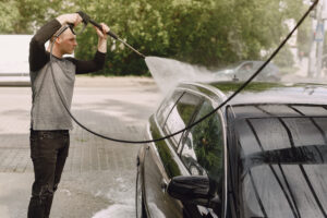 A person uses a pressure washer to clean the roof of a black car outdoors on a paved surface.