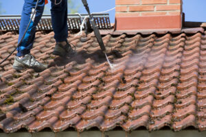 Person cleaning a red tiled roof with a pressure washer, removing moss and dirt buildup; only their lower body and feet are visible.