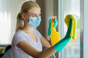 Woman wearing a face mask and green gloves cleaning a window with a yellow spray bottle and yellow cloth.