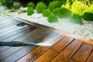 A person uses a power washer to clean a wooden deck, with water visibly removing dirt and grime from the surface. Green shrubs and rocks are in the background.