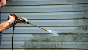 Person using a pressure washer to clean dirt and mildew off the exterior siding of a house; the difference between cleaned and uncleaned areas is visible.