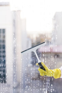 A person wearing a yellow glove cleans a wet window with a squeegee, removing soapy water streaks.