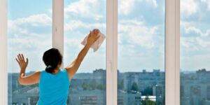 Person in a blue shirt cleaning large windows with a cloth, with a cityscape and blue sky visible outside.