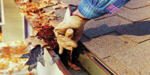 A gloved hand removes leaves from a house gutter filled with autumn foliage next to an asphalt shingle roof.