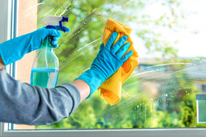 Person wearing blue gloves cleans a window with a yellow cloth and spray bottle, wiping away streaks with an outdoor view in the background.