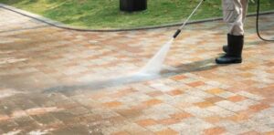 Person using a pressure washer to clean a tiled outdoor surface, with visible difference between cleaned and uncleaned areas.