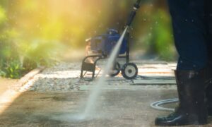 Person using a pressure washer to clean an outdoor walkway, with water spraying from the nozzle and a pressure washer machine in the background.