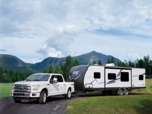 A white pickup truck towing a large travel trailer is parked on a paved road with green fields, trees, and mountains in the background.
