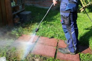 Person using a pressure washer to clean red patio tiles outdoors, with water spray visible and grass surrounding the area.