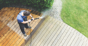 A person uses a pressure washer to clean a wooden deck, revealing a lighter, cleaner section beside a darker, dirtier area.
