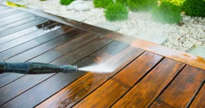 A pressure washer is cleaning a wooden deck, removing dirt and making the wood appear brighter and cleaner. Shrubs and rocks are visible in the background.