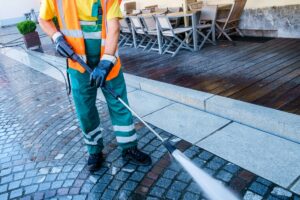 A worker in a safety vest and gloves uses a pressure washer to clean a cobblestone sidewalk next to an outdoor seating area with wooden tables and chairs.