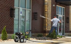A man uses a pressure washer to clean the brick exterior of a commercial building near an accessible parking spot.