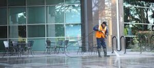 A worker in an orange safety vest uses a pressure washer to clean the outdoor area in front of a glass building with empty tables and chairs.