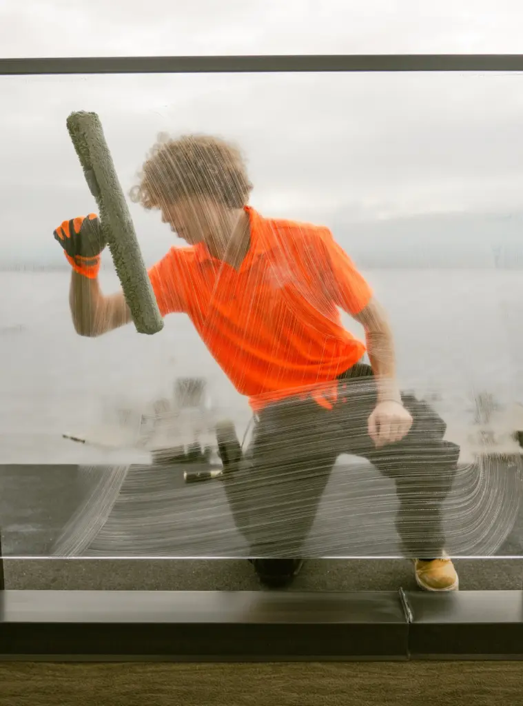 Professional window cleaner in bright orange shirt scrubs glass railing overlooking the lake in Lake Country BC during overcast day.