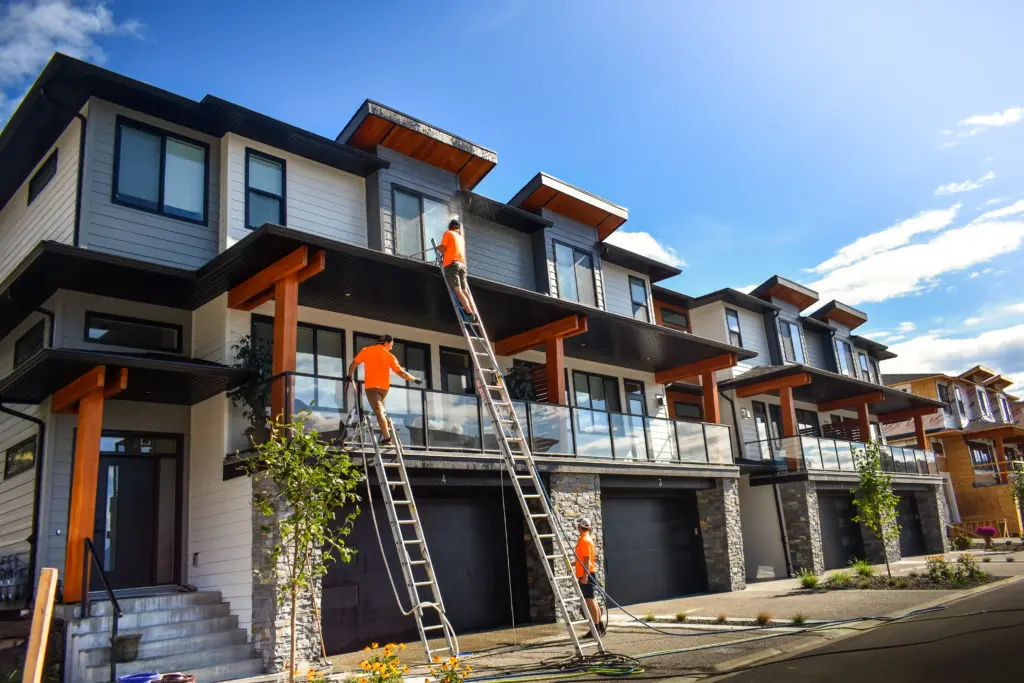 Three workers in orange shirts pressure washing the upper floors and windows of modern townhomes in Lake Country, BC using ladders and hoses.