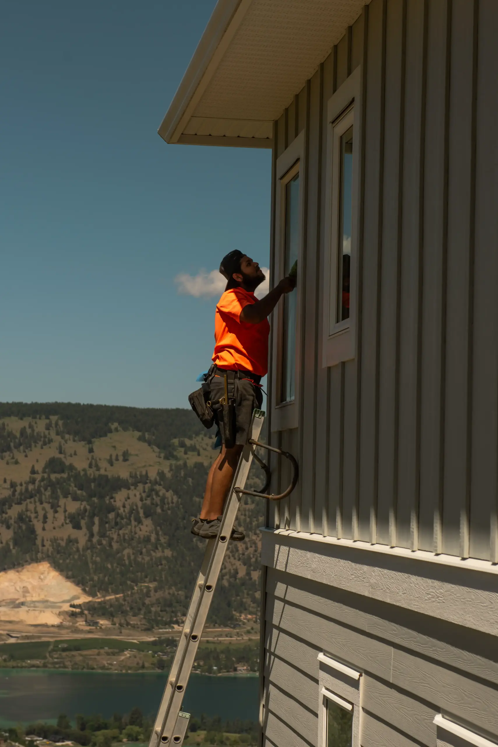 Professional window cleaner in orange shirt on ladder cleaning second-story window with Lake Country BC lake and mountain views in background.