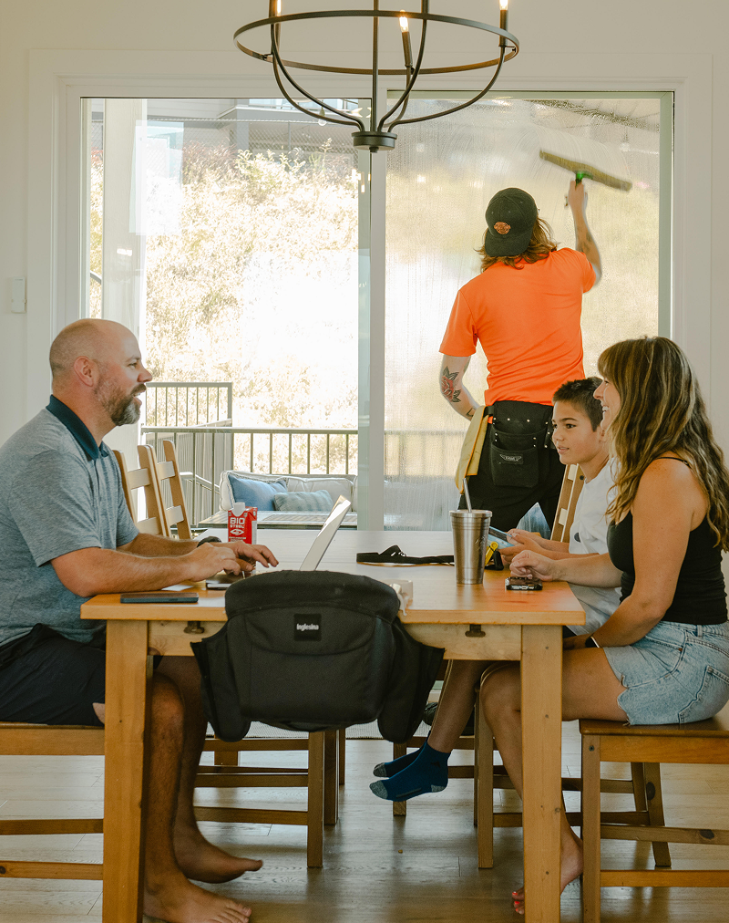 A man cleans a large glass door while three people sit at a dining table, two working on laptops and one watching.