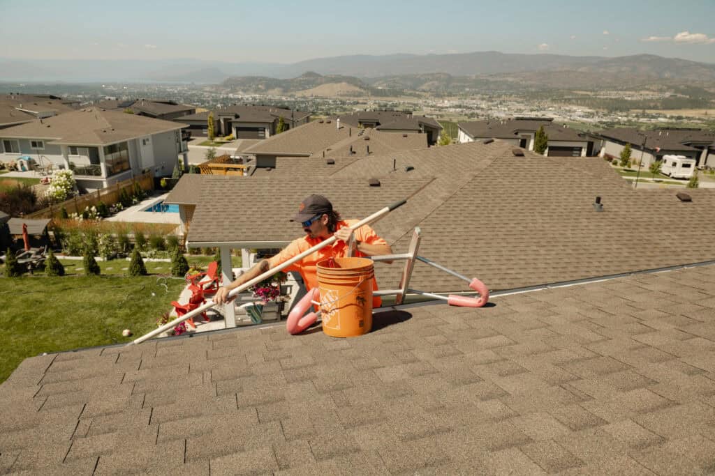 Technician cleaning gutters on a rooftop overlooking a Kelowna neighbourhood